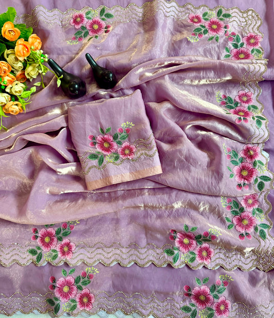 A lavender purple embroidered fabric set with pink floral motifs, green leaves, and gold zari borders, arranged in a styled flat lay with a matching folded cloth, two black decorative objects, and a small bouquet of orange and yellow flowers.
